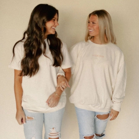 Two women wearing matching cream sweatshirts and light blue jeans against a beige background.