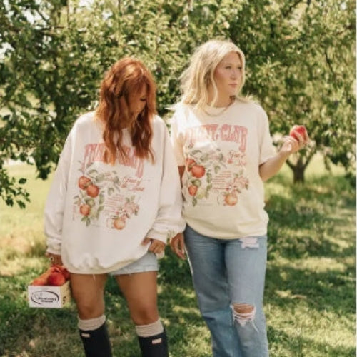 Two women in an apple orchard wearing matching sweatshirts with apple designs.