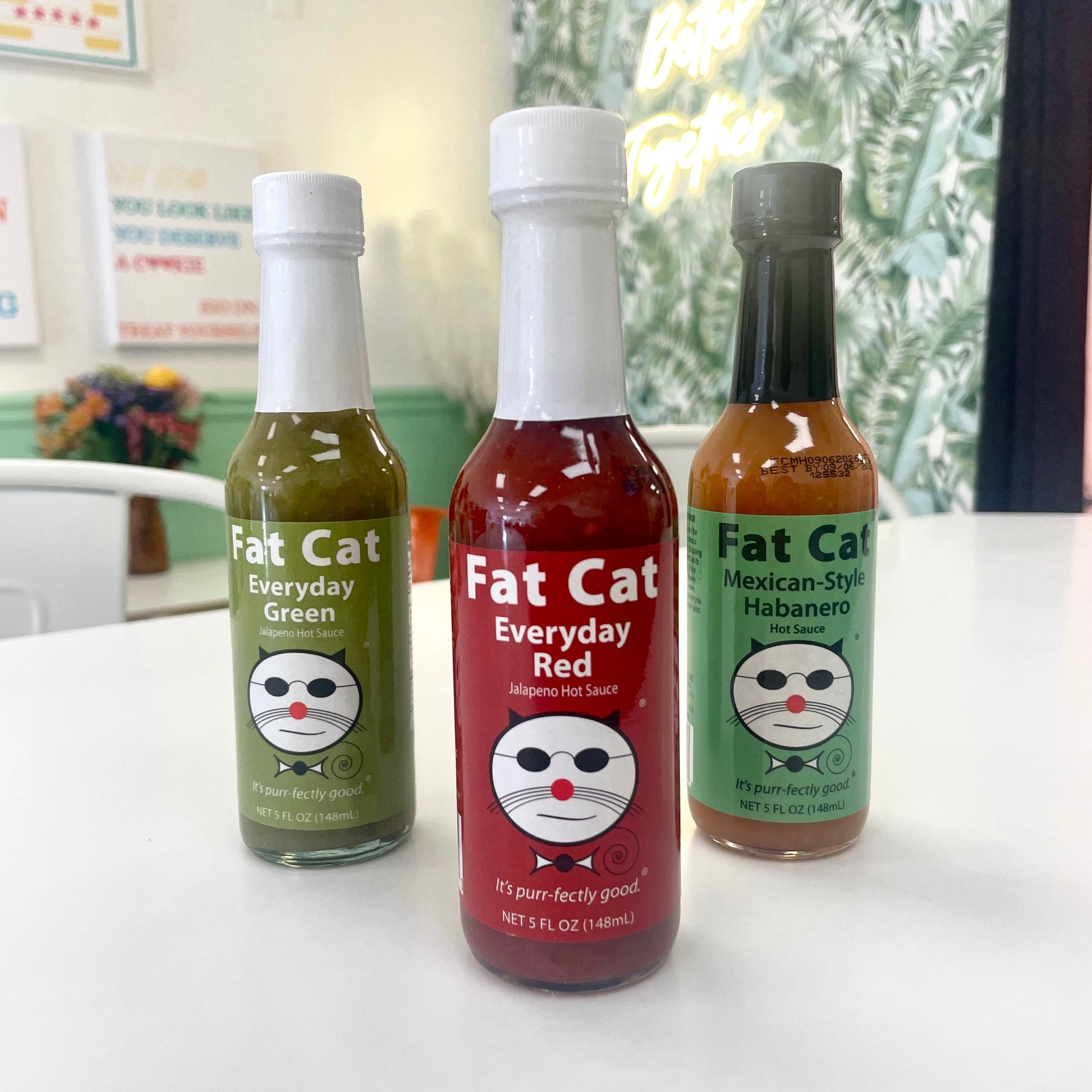 Three bottles of Fat Cat hot sauce on a table with a blurred indoor background