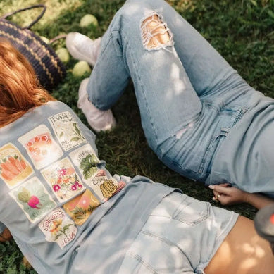 Person sitting on grass wearing ripped jeans and denim shorts with a basket of flowers.