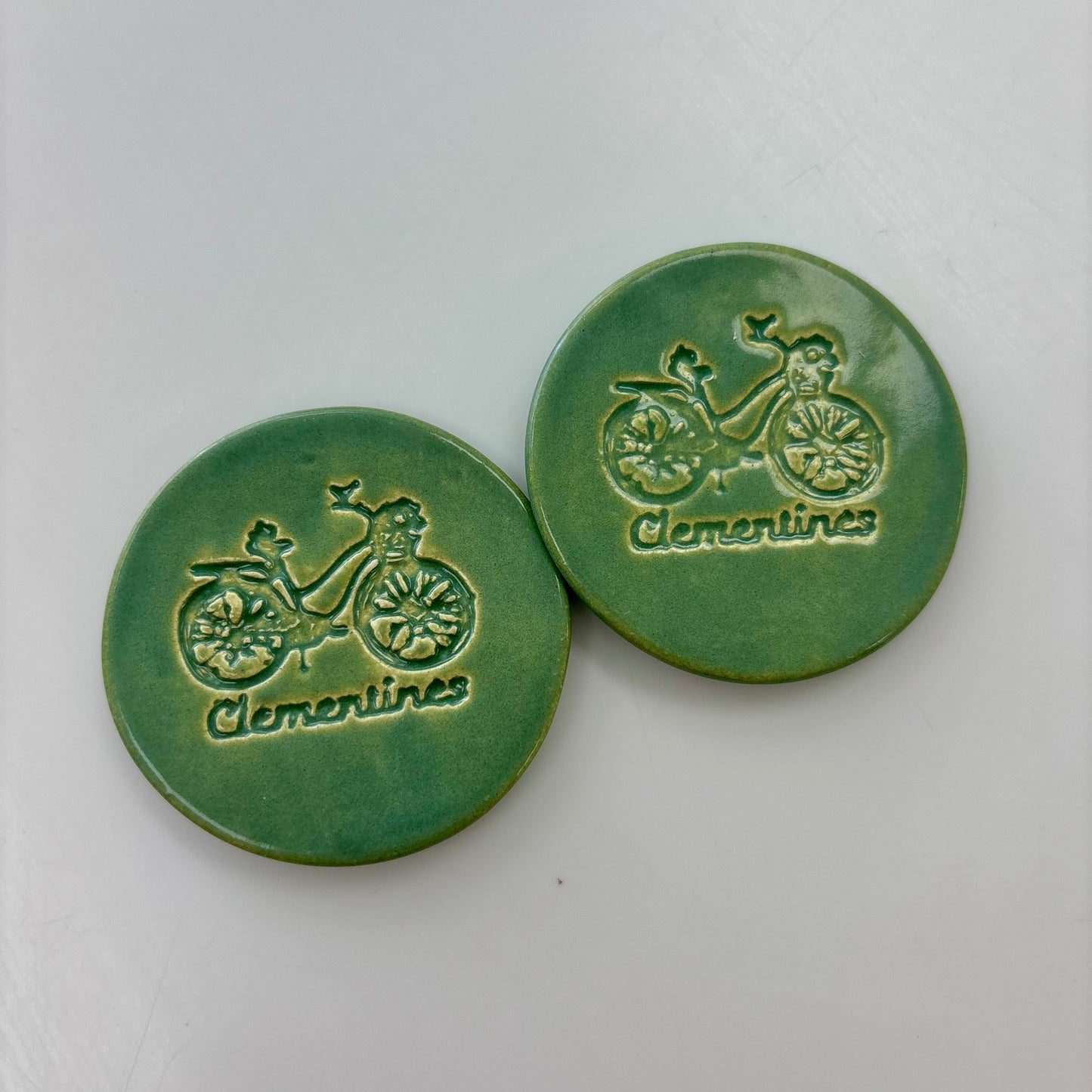 Two green ceramic buttons with bicycle design and 'Clementines' text on a white background
