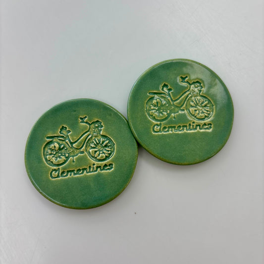 Two green ceramic buttons with bicycle design and 'Clementines' text on a white background