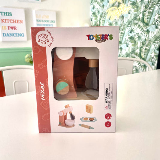 Toy mixer in packaging on a table with a floral-patterned curtain in the background