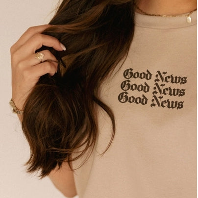 Person wearing a beige t-shirt with 'Good News' text, holding hair.