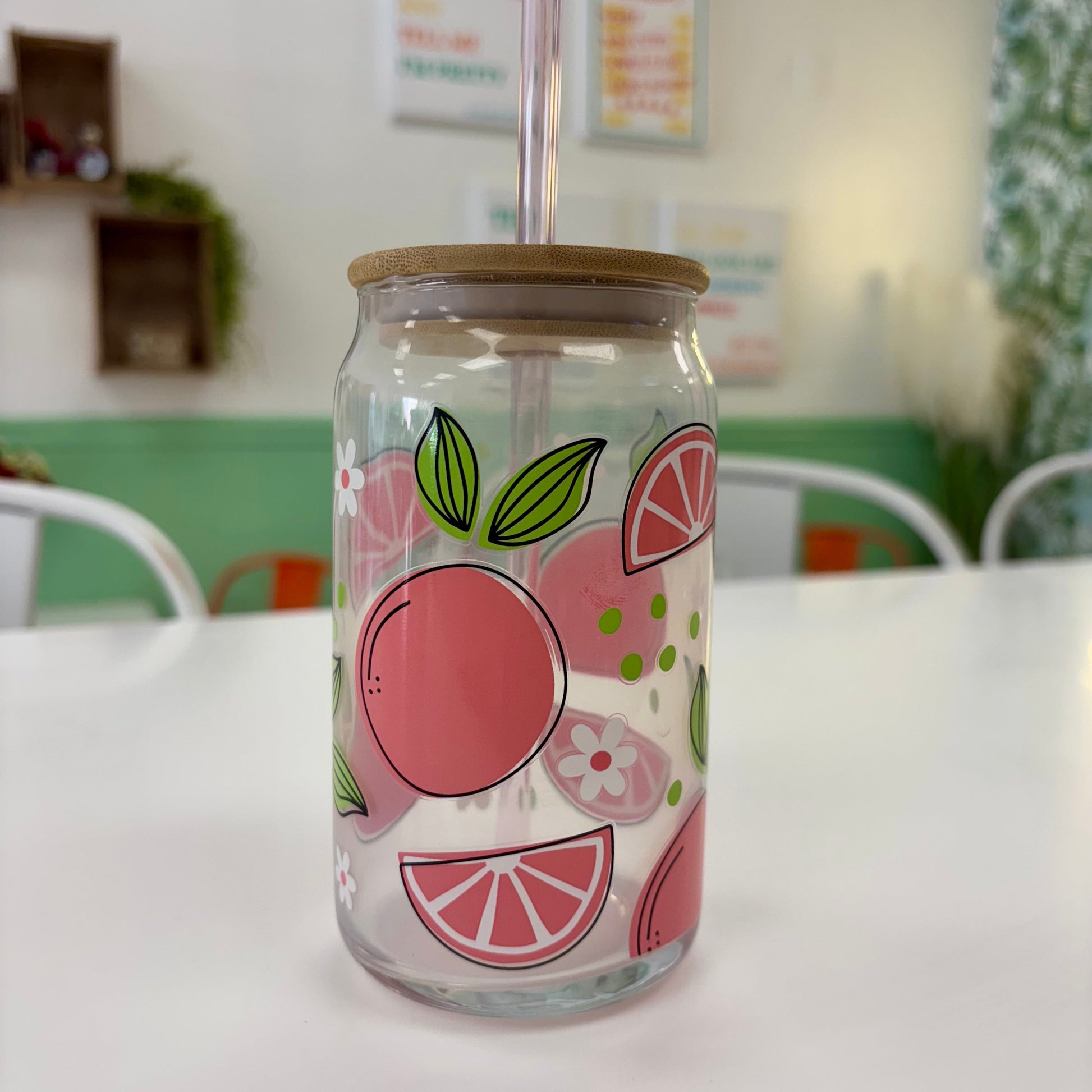 Glass jar with a wooden lid and straw, featuring fruit designs on a white table.